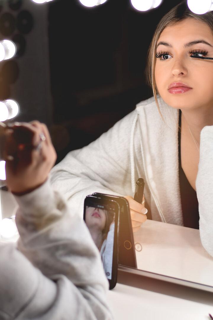 Junge Frau trägt vor einem beleuchteten Spiegel Mascara auf und filmt sich dabei mit ihrem Smartphone.