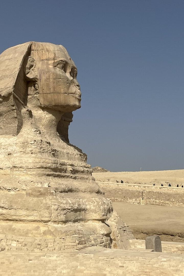 Die Große Sphinx von Gizeh mit Löwenkörper und Menschenkopf steht in einer sandigen Wüstenlandschaft.