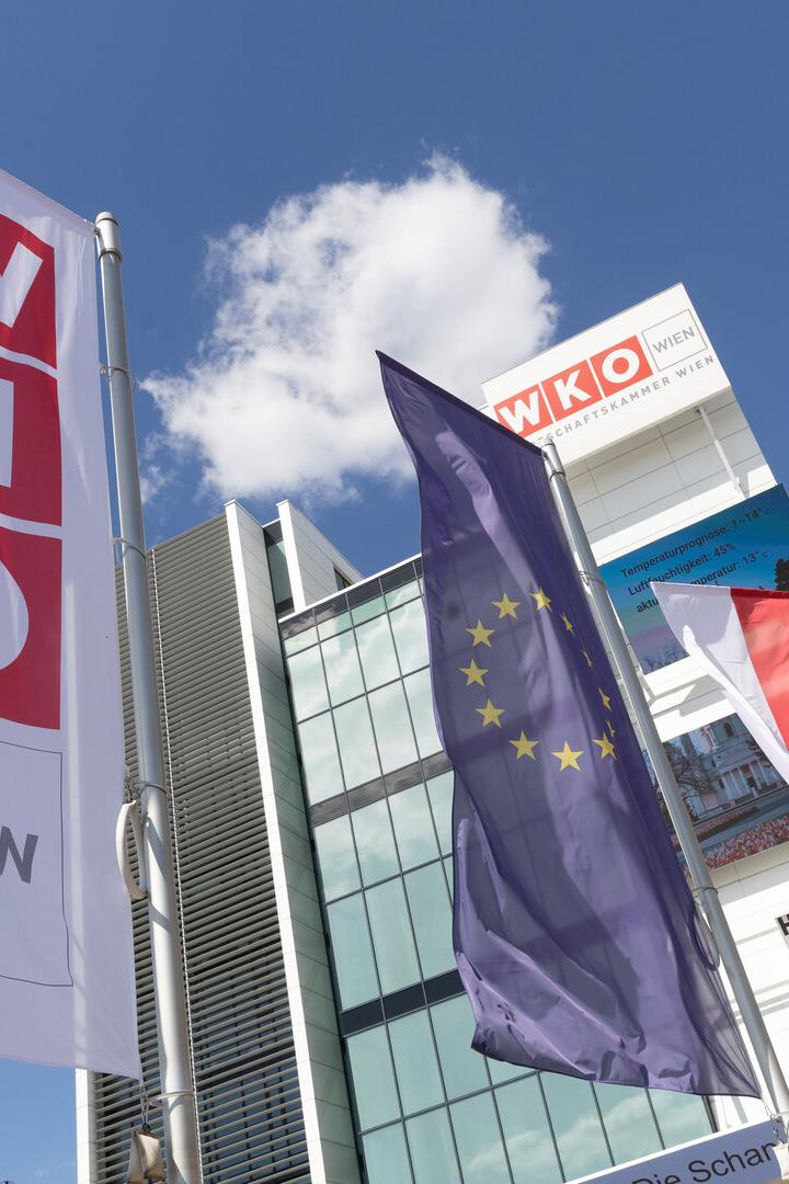Mehrere Fahnen, darunter die der Wirtschaftskammer Wien, der EU und Österreichs, wehen vor einem modernen Gebäude bei blauem Himmel.