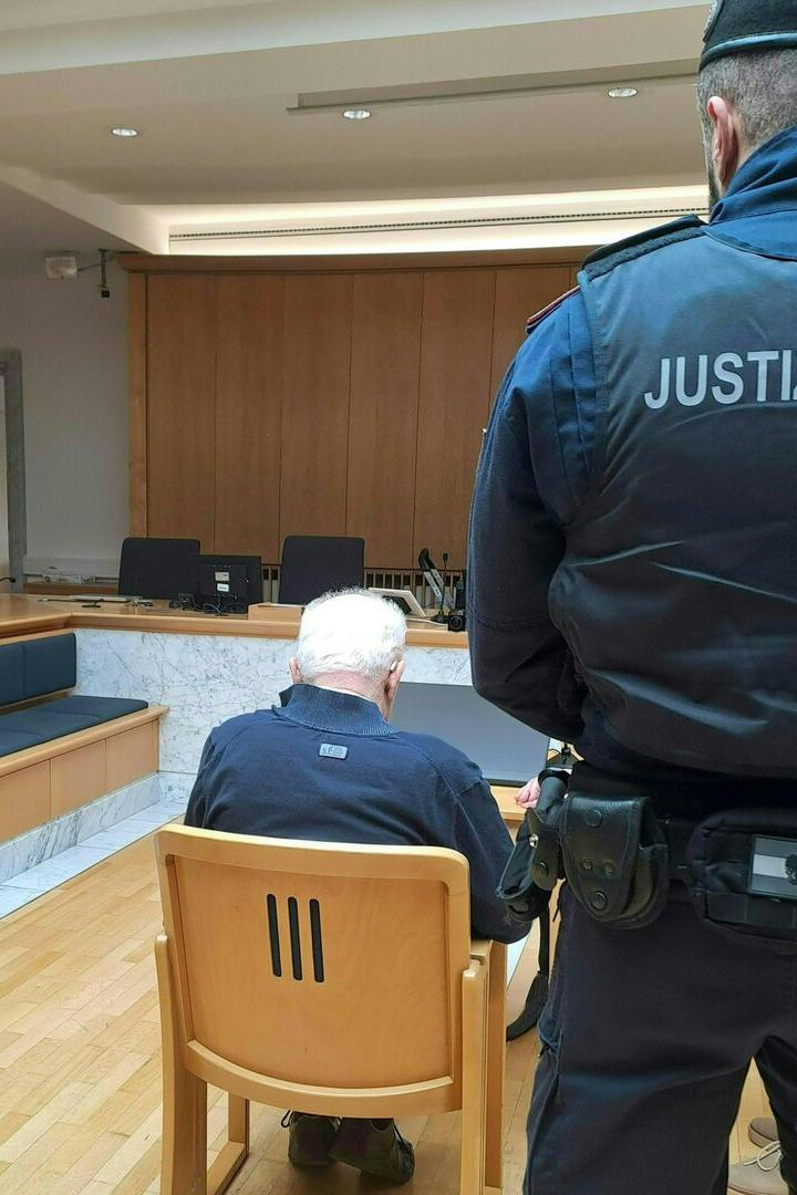 Ein Mann mit weißem Haar sitzt in einem Gerichtssaal, bewacht von einem Justizbeamten.