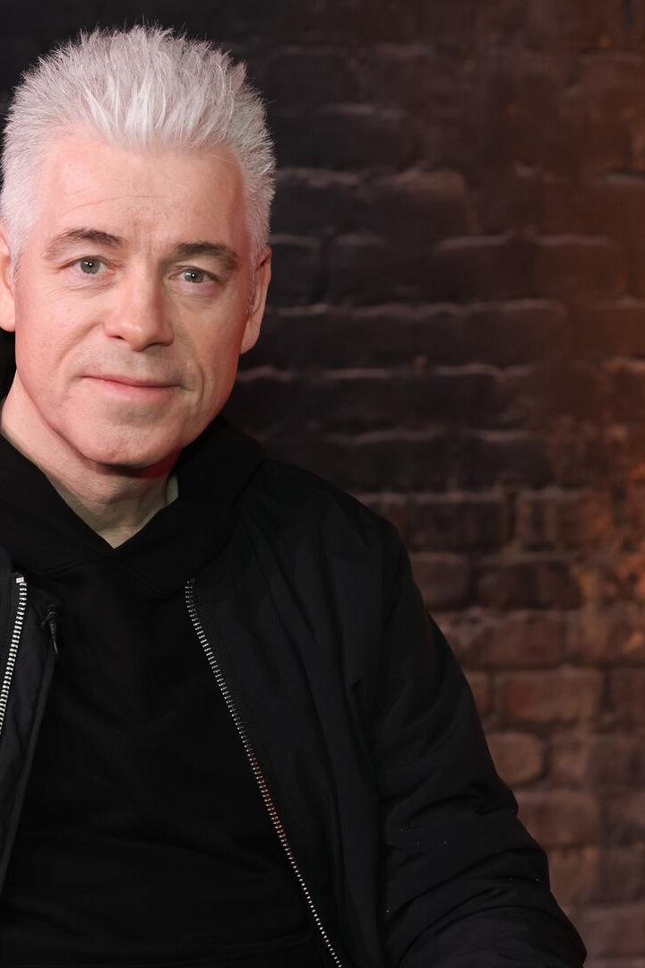 Michael Mittermeier trägt eine schwarze Jacke und sitzt vor einer Backsteinwand mit warmem Licht.