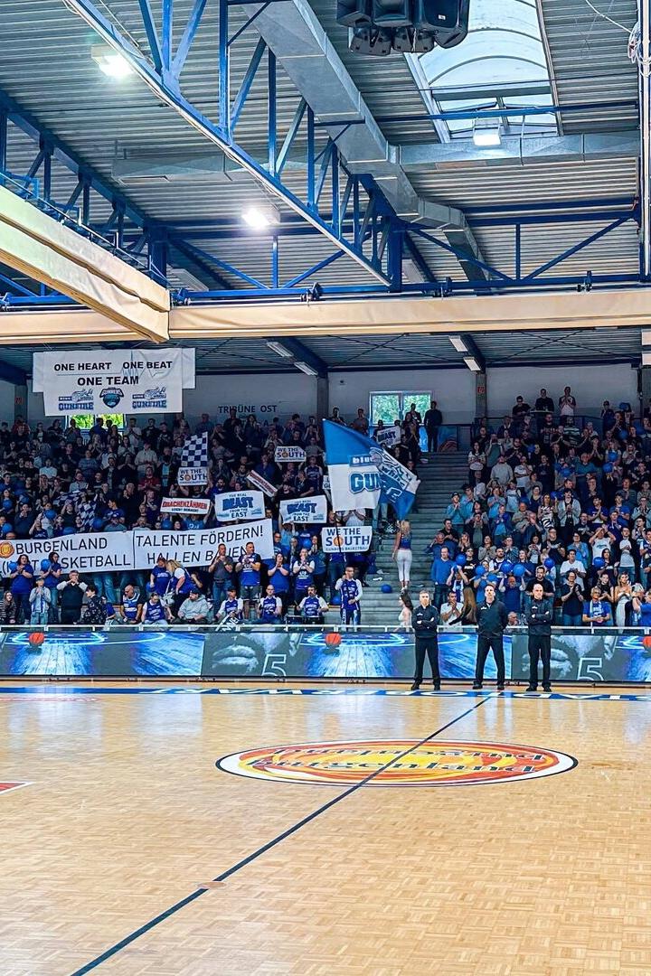 Viele Fans in blauer Kleidung und mit Bannern feuern ein Basketballteam in einer vollen Sporthalle an.