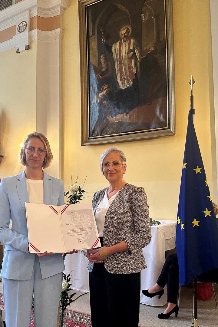 Zwei Frauen halten eine Urkunde vor Flaggen Österreichs und der EU in einem Raum.