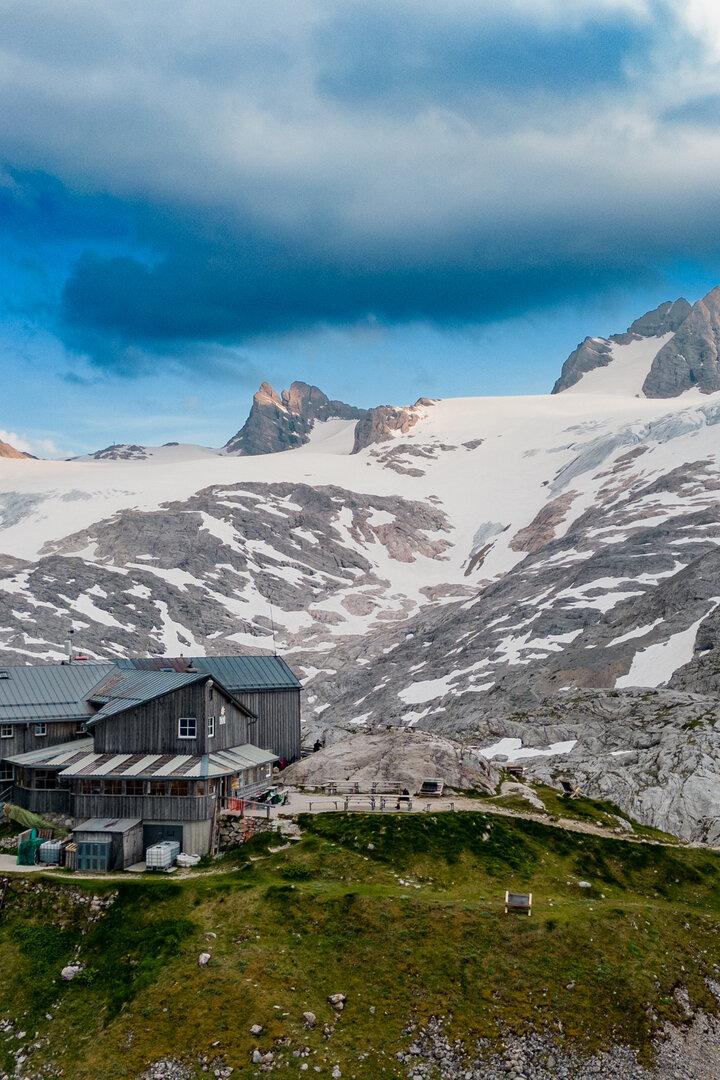 Berghütte mit mehreren Gebäuden auf grünem Hang, umgeben von schneebedeckten Felsen und hohen Bergen unter bewölktem Himmel.