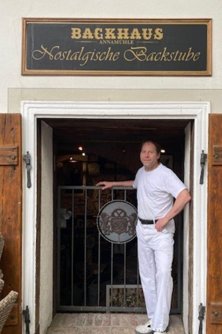 Ein Mann in weißer Kleidung steht in der Tür einer traditionellen Bäckerei mit Holzläden und nostalgischem Schild.