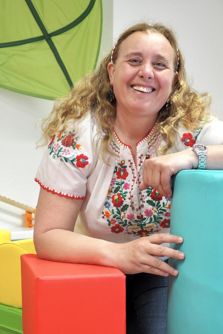 Eine lachende Frau mit lockigem Haar lehnt an bunten Schaumstoffblöcken in einem freundlich eingerichteten Raum.