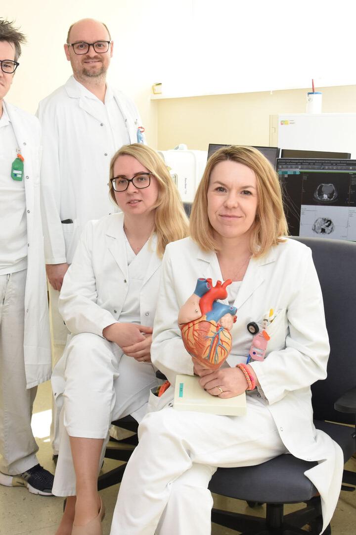 Vier Personen in weißen Kitteln, zwei sitzen, zwei stehen, eine hält ein anatomisches Herzmodell, im Hintergrund Computer mit medizinischen Bildern.
