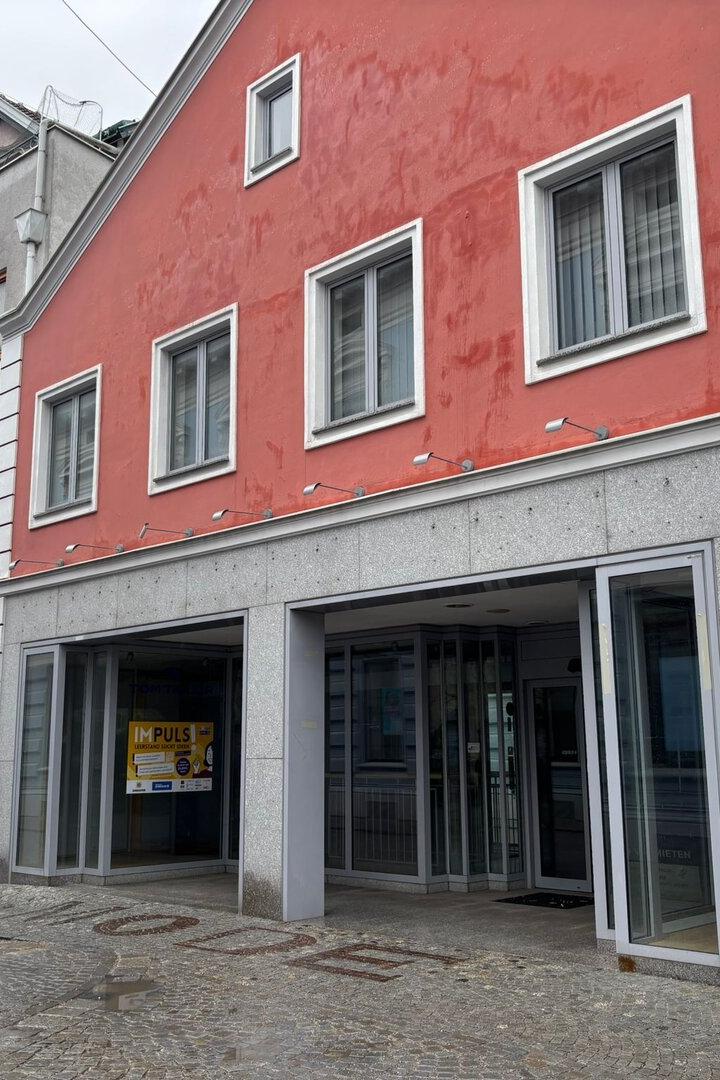 Mehrstöckiges Gebäude mit roter Fassade, mehreren Fenstern und einem offenen Eingang an einer gepflasterten Straße.