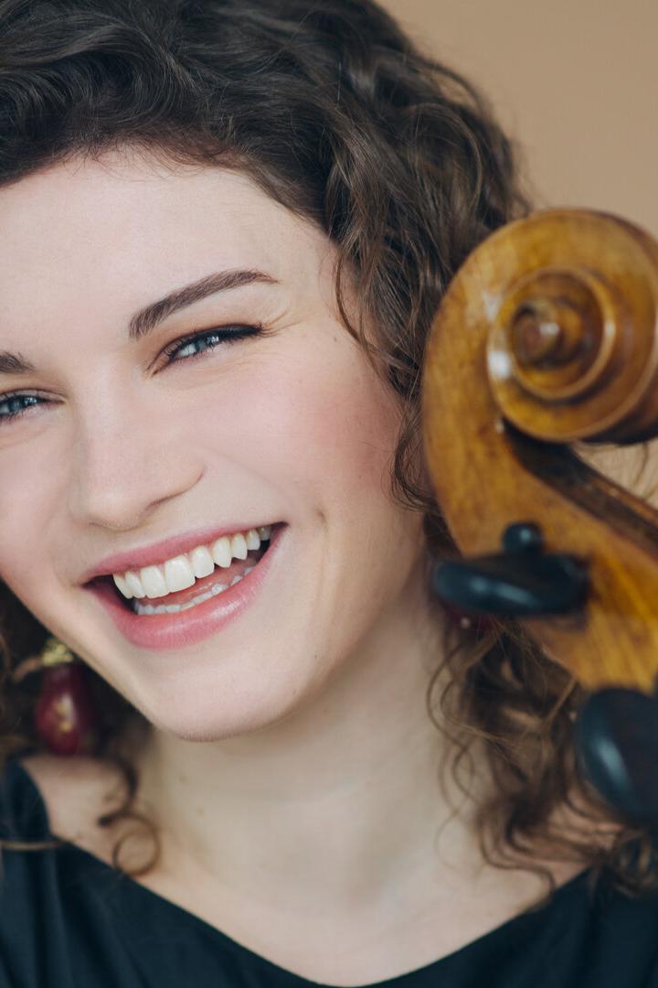 Junge Frau mit lockigen Haaren lächelt und hält ein Streichinstrument mit sichtbarem Wirbel neben ihrem Gesicht.