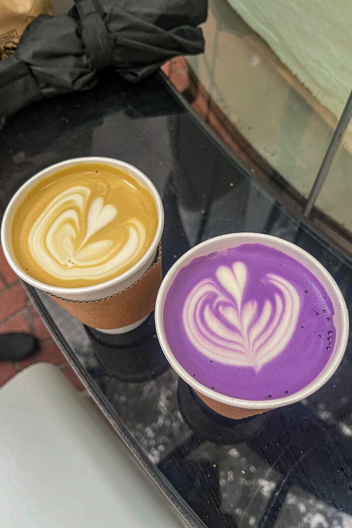 Zwei Becher mit Latte Art, einer mit gelbem und einer mit lila Getränk, stehen nebeneinander auf einem Tisch.