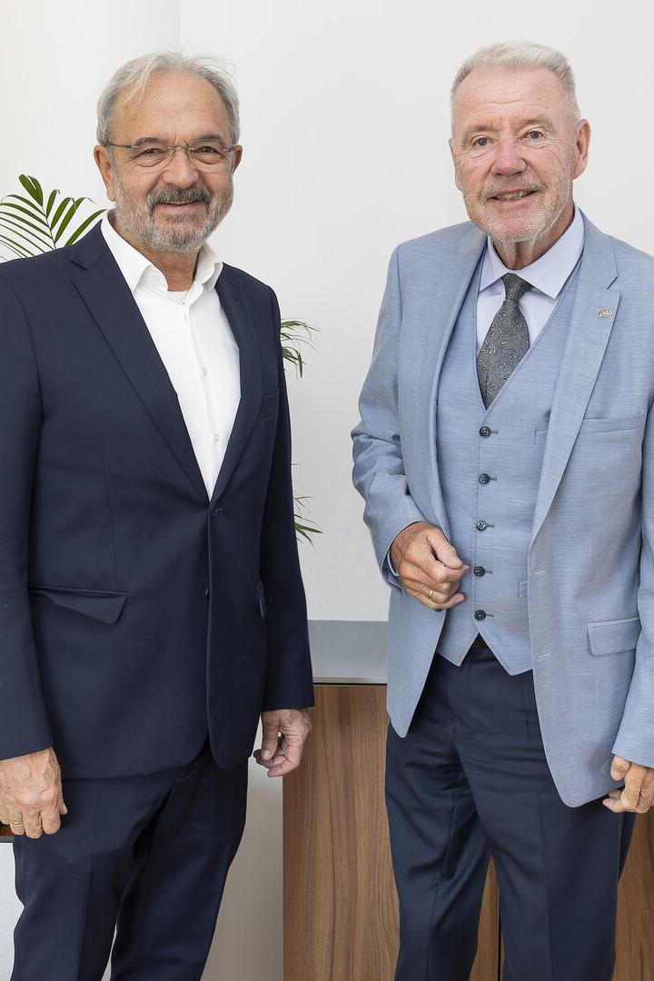 Zwei ältere Männer in Anzügen stehen nebeneinander vor einer weißen Wand mit einer Pflanze im Hintergrund.