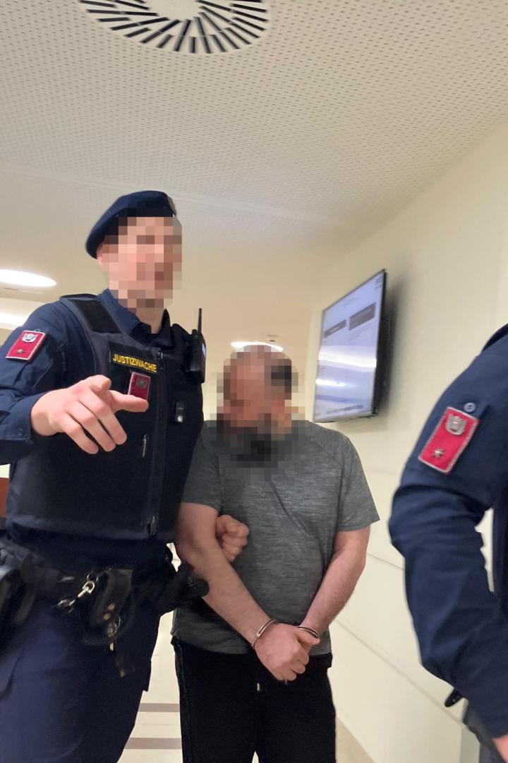 Zwei Justizwachebeamte führen einen gefesselten Mann durch einen Gerichtsflur.