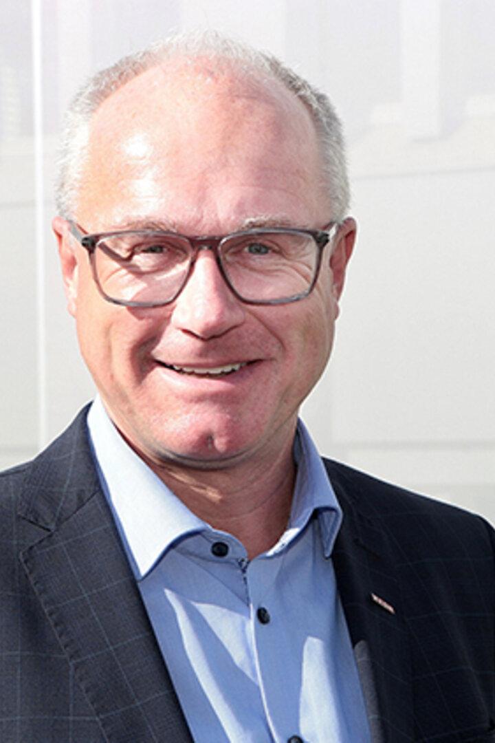 Ein älterer Mann mit Brille und Anzug lächelt vor einem hellen, unscharfen Hintergrund.