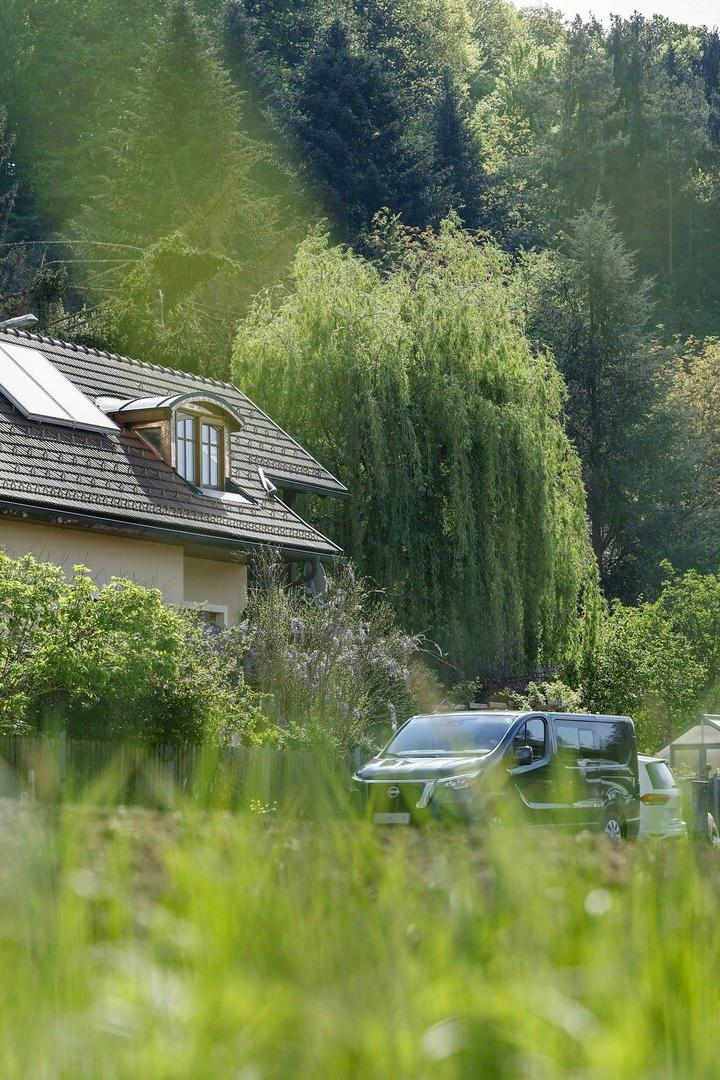 Ein Haus mit Dachfenstern steht vor einem bewaldeten Hügel, davor parken Autos.