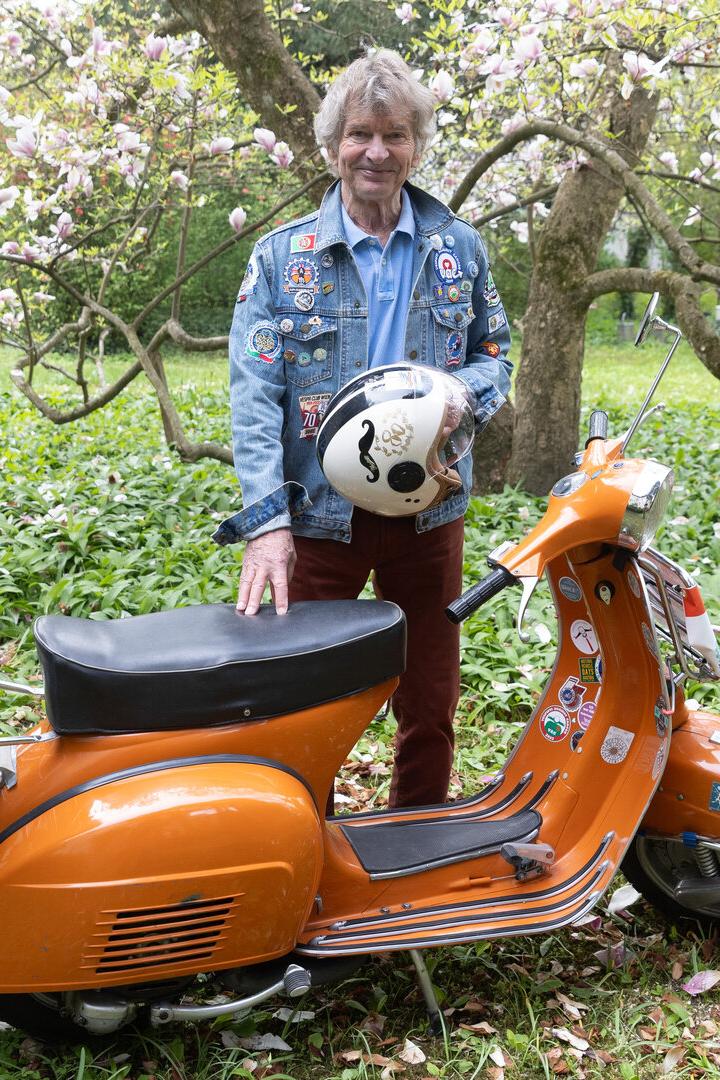 Ein älterer Mann steht im Garten neben einem orangefarbenen Motorroller und hält einen Helm in der Hand.