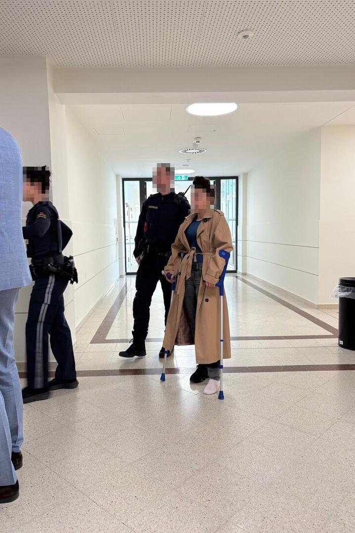 Eine Person mit Krücken und beigem Mantel steht neben Polizisten in einem hellen Flur.