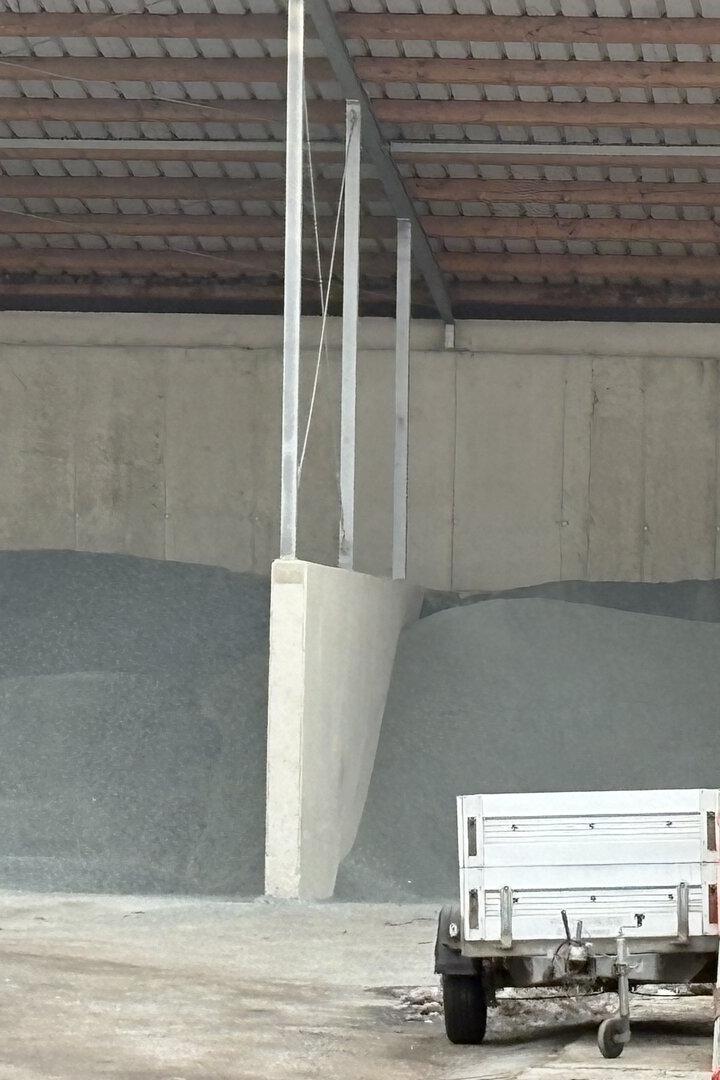 Zwei Haufen Streusalz oder Splitt unter einem Dach, daneben ein Kommunalfahrzeug mit Anhänger und Müllcontainer.