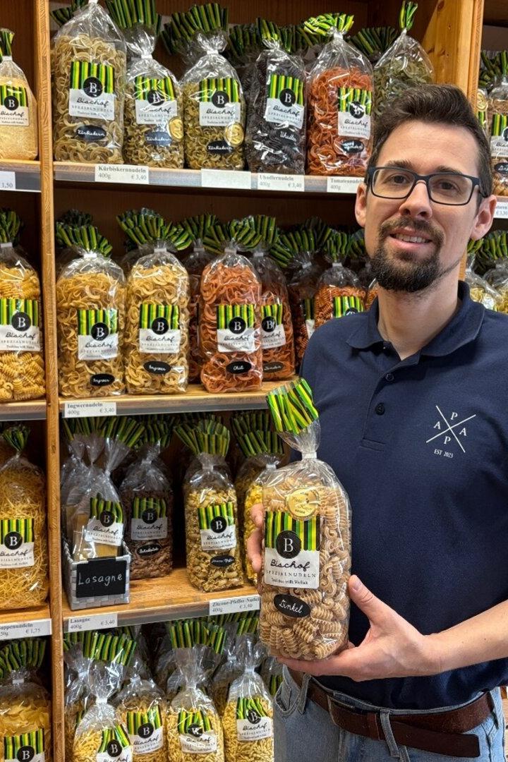 Ein Mann mit Brille steht vor Regalen voller verschiedener Nudelsorten und hält eine Packung Nudeln in der Hand.