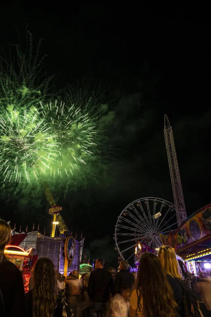 Menschenmenge auf einem Jahrmarkt bei Nacht, bunte Fahrgeschäfte und grünes Feuerwerk am Himmel.
