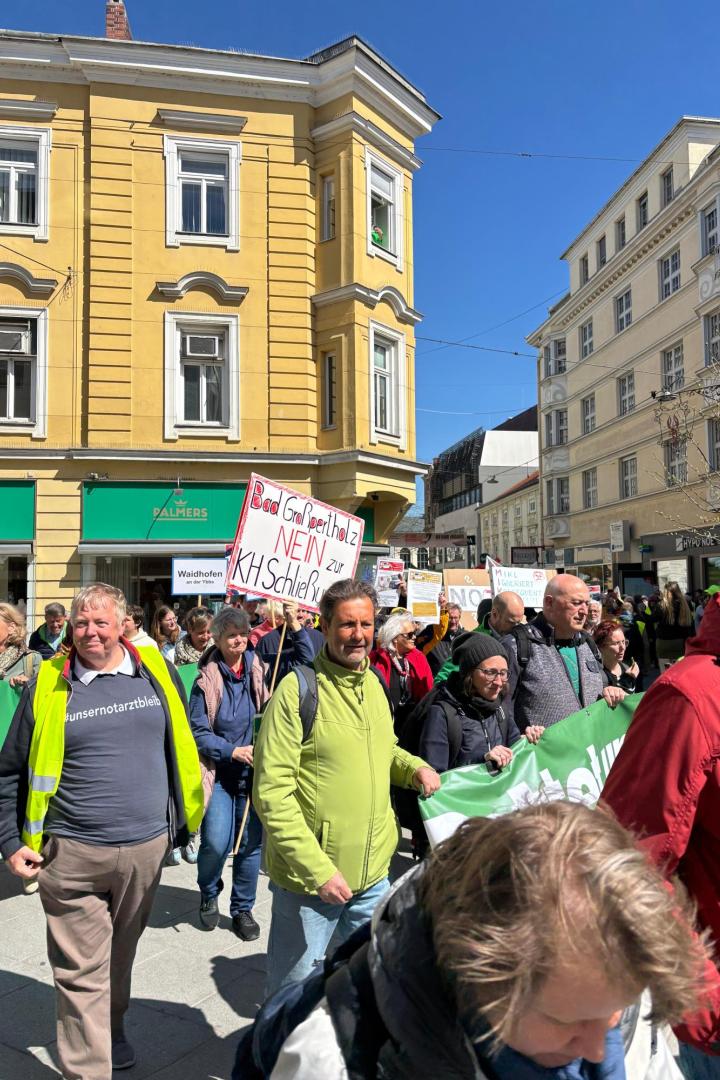 Eine Gruppe von Menschen demonstriert mit Transparenten und Schildern auf einer sonnigen Straße vor gelben Gebäuden.