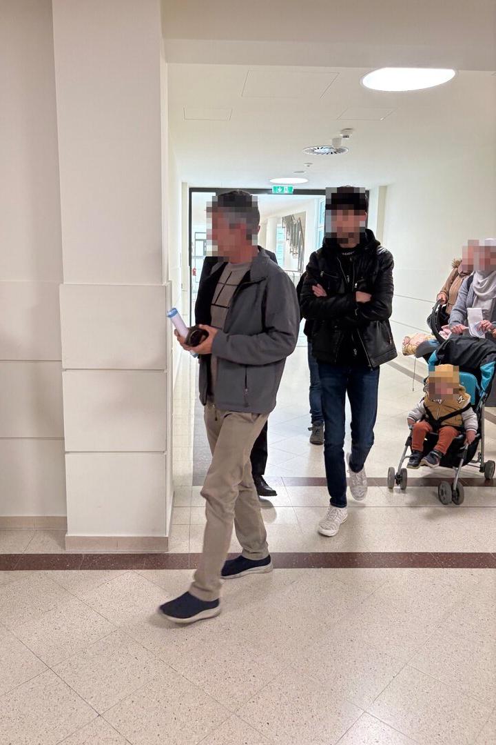 Mehrere Personen gehen durch einen hellen Flur, darunter ein Mann mit Dokumenten und ein Kind im Buggy.