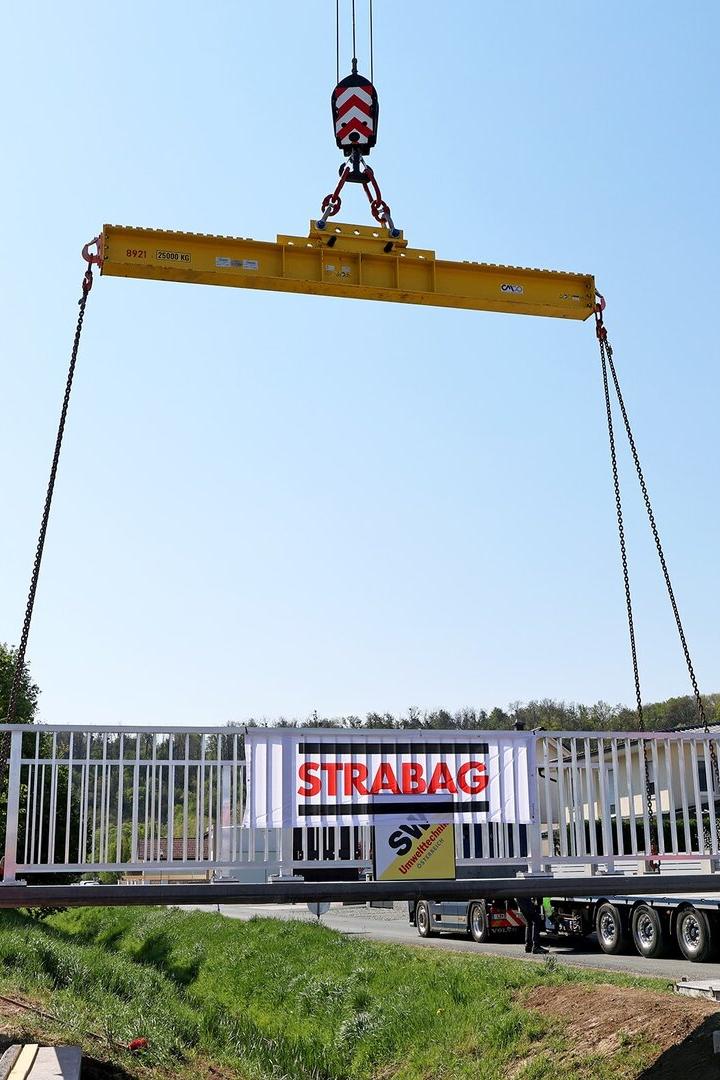Ein Kran hebt eine weiße Brücke mit STRABAG-Logo, während zwei Bauarbeiter in Warnkleidung den Bereich sichern.