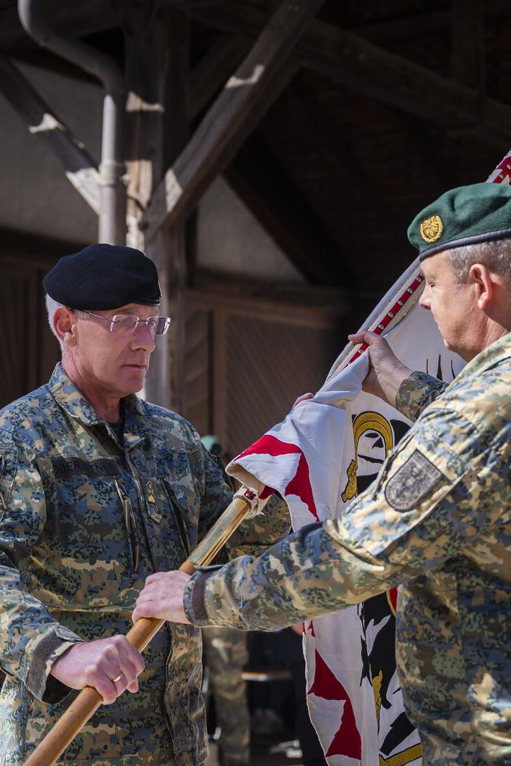 Zwei Soldaten in Uniform übergeben eine Fahne vor einer Gruppe von Personen.