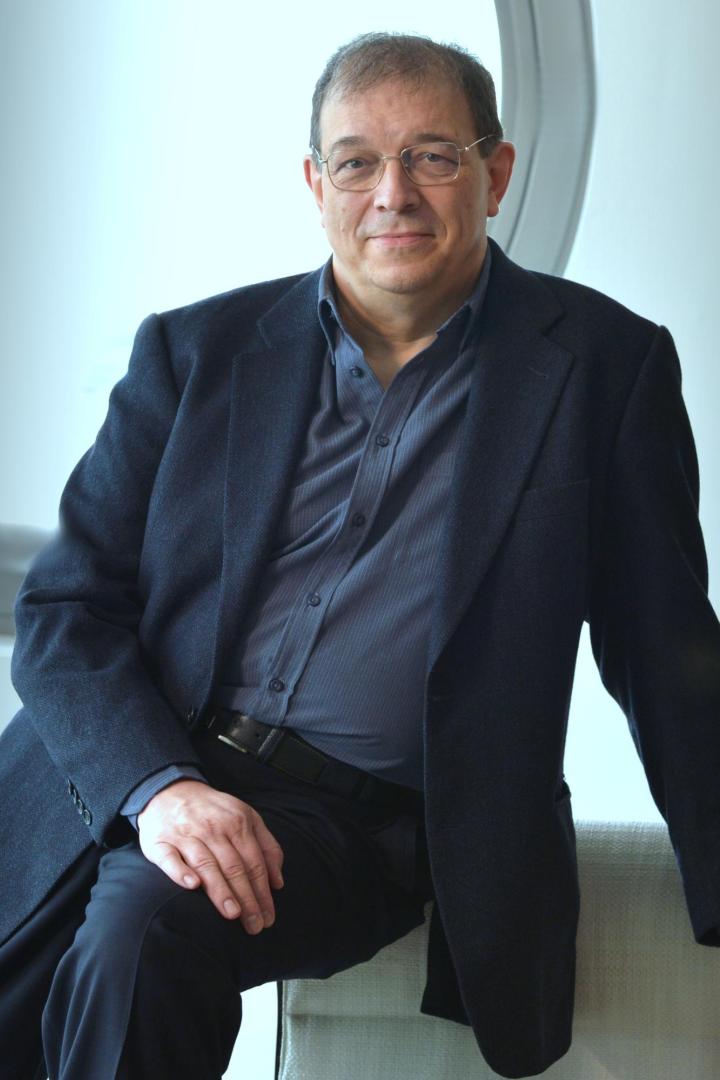 Ein Mann mit Brille und dunklem Sakko sitzt entspannt auf einem Stuhl vor einem hellen, modernen Hintergrund.