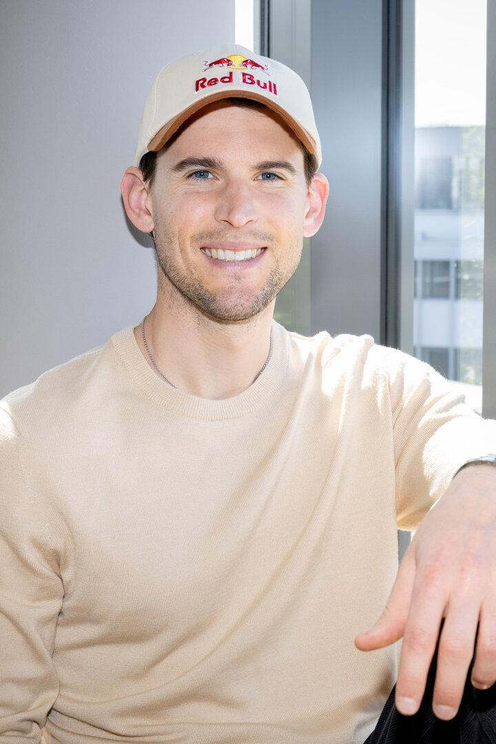 Ein junger Mann mit beigem Pullover und Red Bull-Kappe sitzt lächelnd vor einem Fenster mit modernem Rahmen.