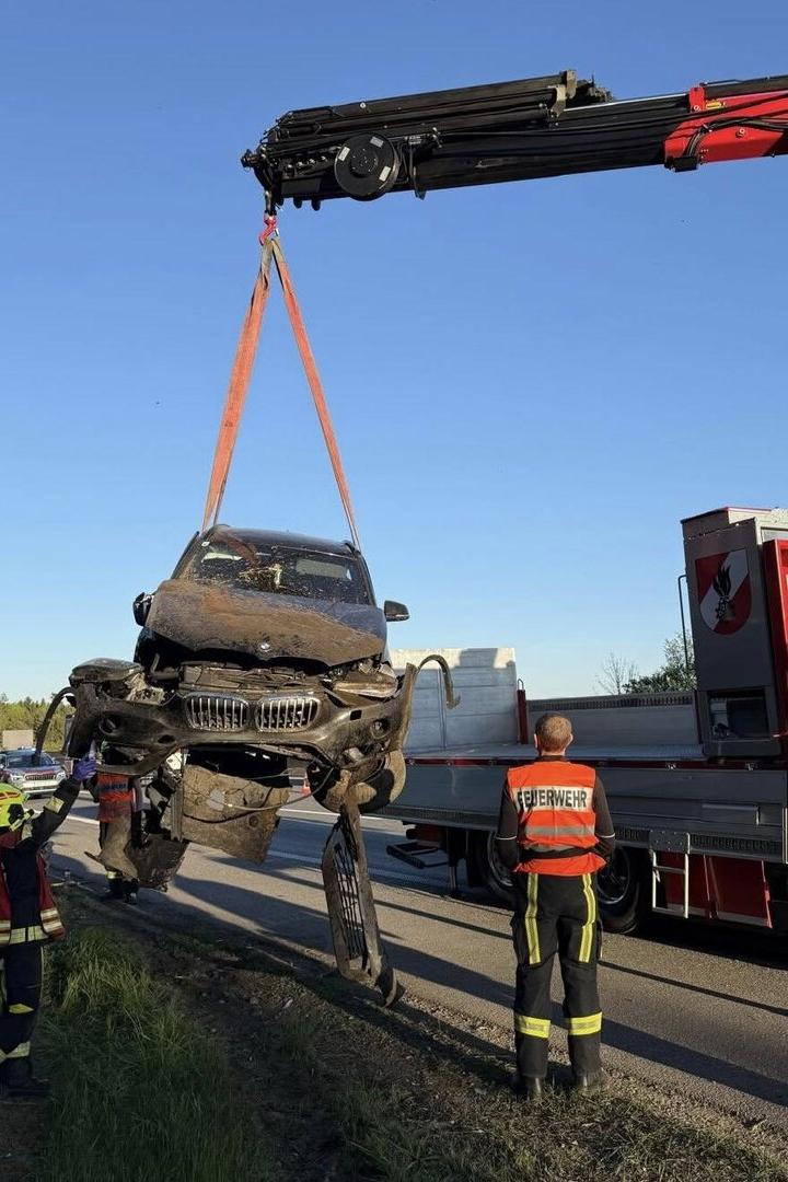 Ein stark beschädigtes Auto wird von einem Kran angehoben, während Feuerwehrleute daneben stehen.