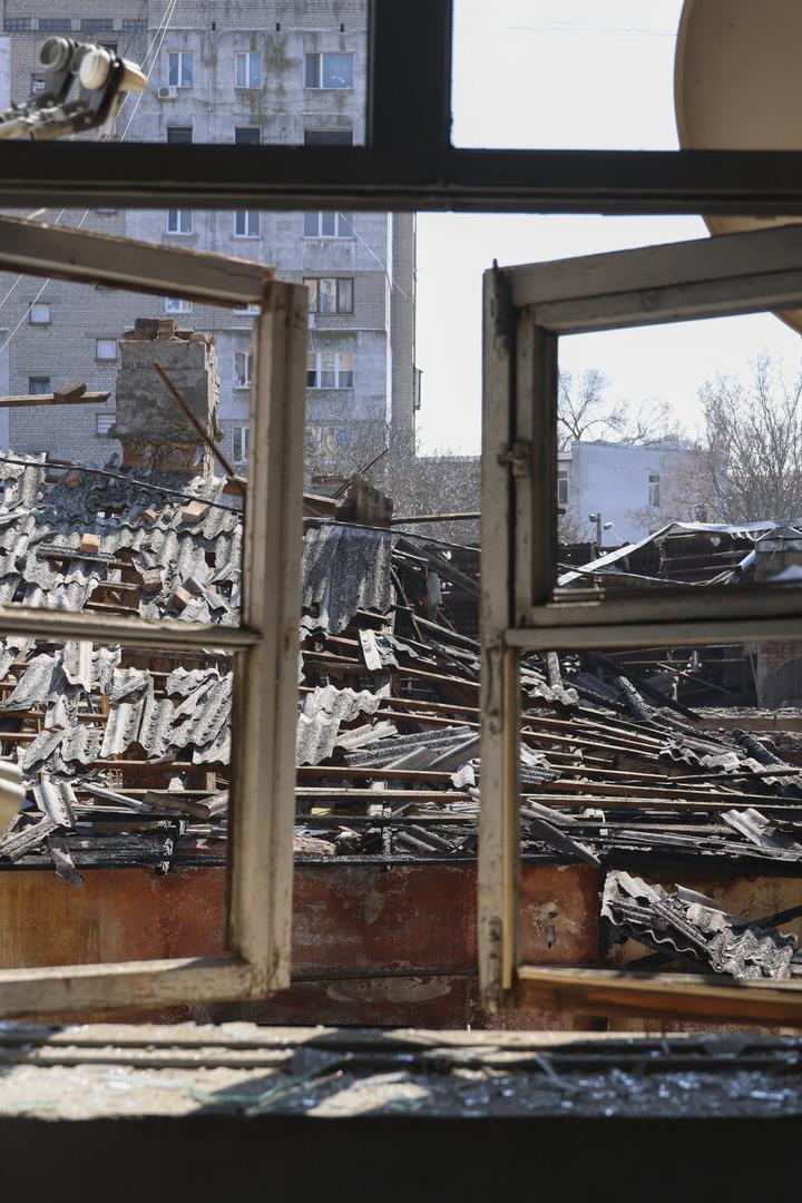 Durch ein geöffnetes Fenster sieht man ein zerstörtes Dach und umliegende Gebäude.