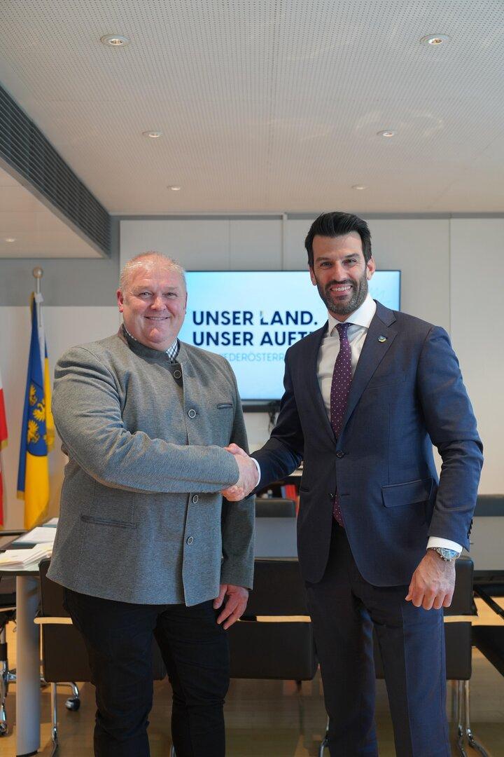 Zwei Männer schütteln sich vor einem Bildschirm mit der Aufschrift "Unser Land. Unser Auftrag.".