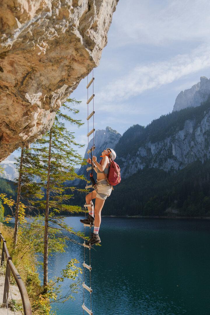 Eine Frau mit Rucksack klettert eine Leiter an einer Felswand über einem See hinauf.