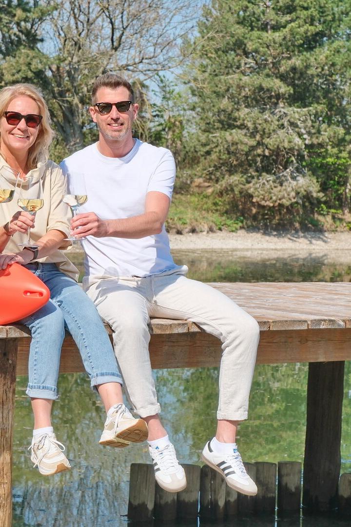 Drei Personen sitzen lachend auf einem Steg am Wasser, stoßen mit Gläsern an und tragen Sonnenbrillen.