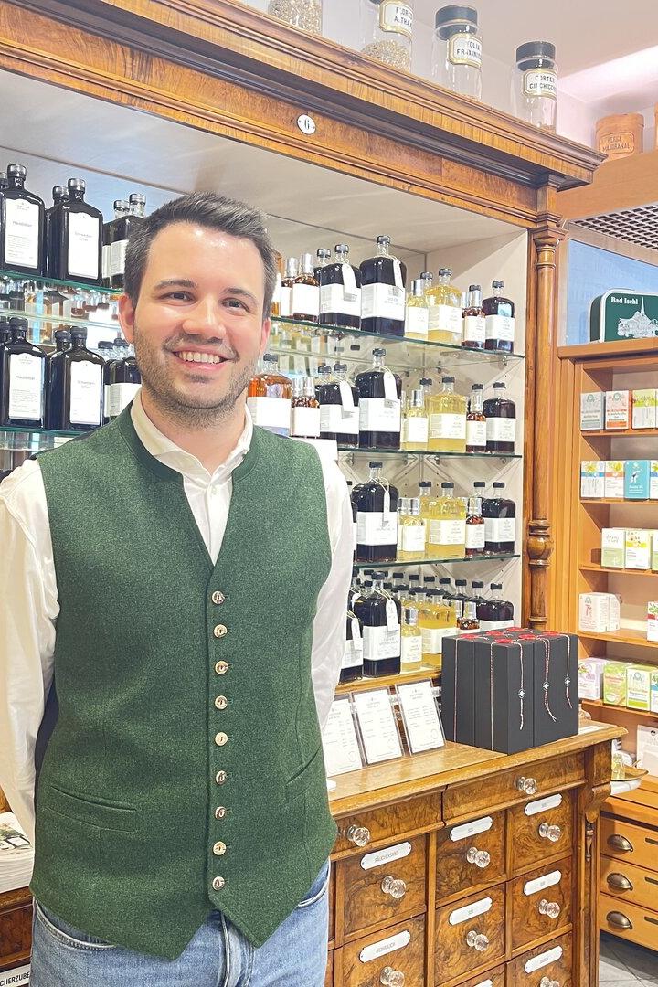 Ein Mann mit grünem Gilet steht lächelnd in einer traditionellen Apotheke vor Regalen mit Flaschen und Produkten.