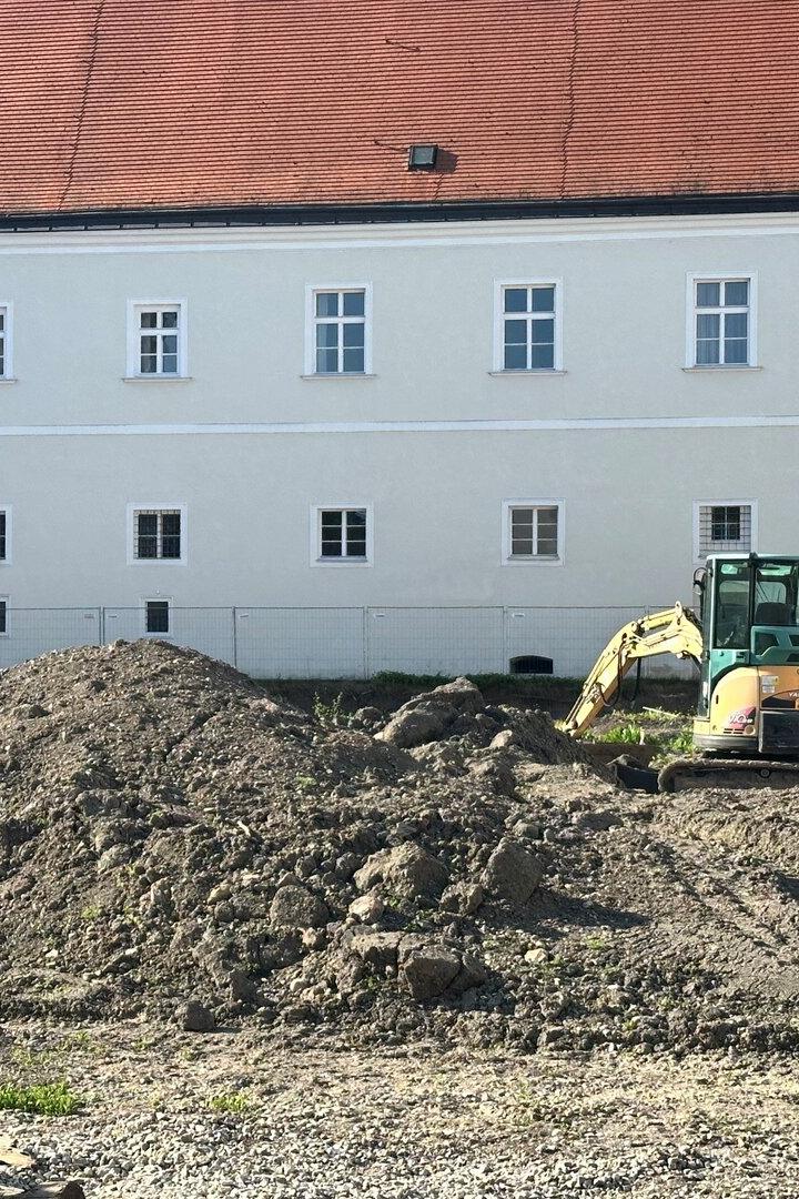 Ein Bagger steht auf einer Baustelle vor einem großen, weißen Gebäude mit rotem Dach.