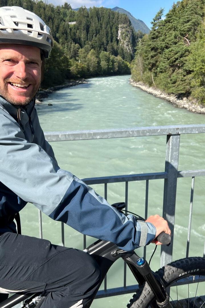 Ein Mann mit Helm und Rucksack fährt auf einem Fahrrad über eine Brücke mit Fluss und Wald im Hintergrund.
