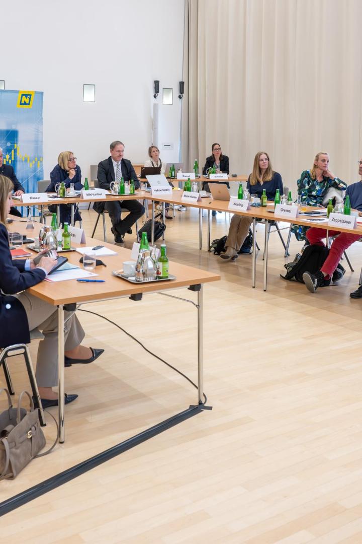 Viele Menschen sitzen in einem hell erleuchteten Raum vor einem Aufsteher des Wirtschaftslandes NÖ um einen Tisch.
