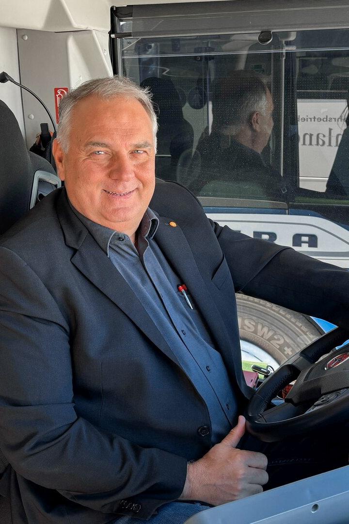 Ein lächelnder Mann im Anzug sitzt am Steuer eines Busses.