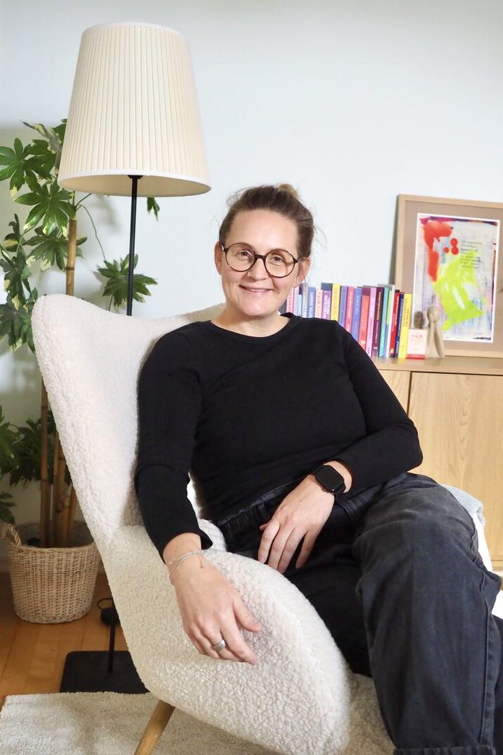 Eine Frau mit Brille sitzt entspannt in einem Sessel, im Hintergrund stehen eine Stehlampe, Pflanzen und ein Regal mit Büchern.