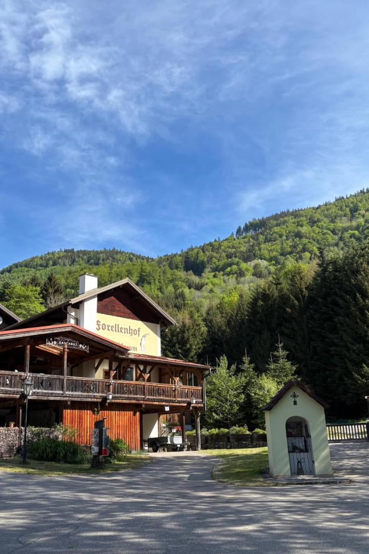 Ein Gasthaus mit Holzveranda steht vor bewaldeten Hügeln, daneben eine kleine Kapelle und umgeben von grünen Bäumen.