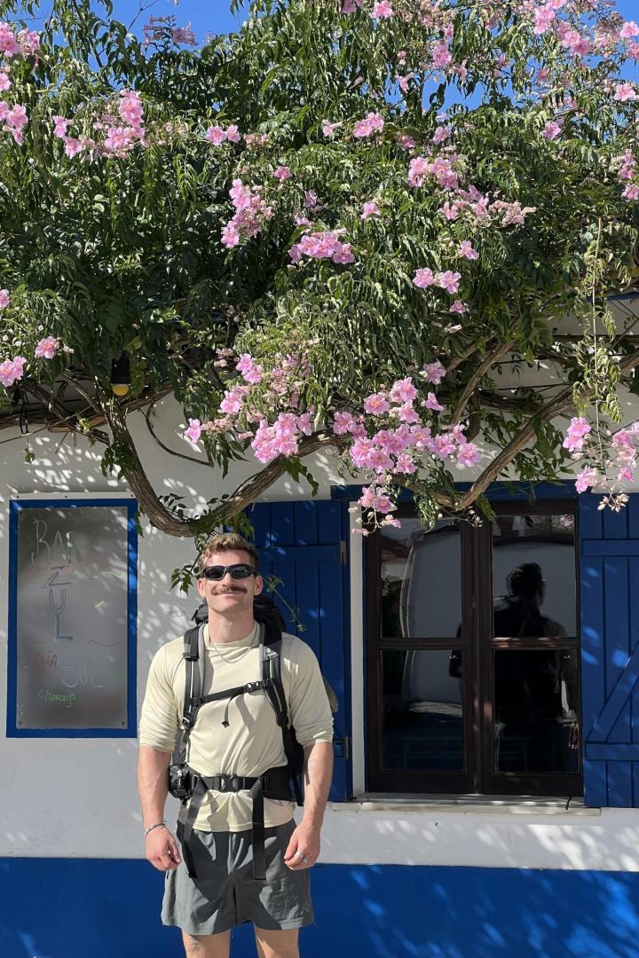 Ein junger Mann steht unter einem blühenden Baum, mit Rucksack, kurzer Hose und Sonnenbrille.