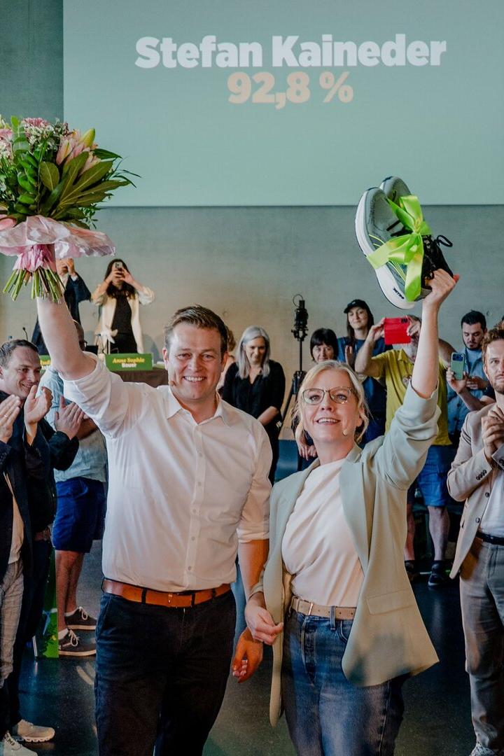 Zwei Personen stehen jubelnd inmitten applaudierender Menschen, eine hält Blumen, die andere ein Paar Sportschuhe hoch.