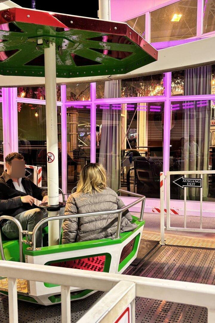 Zwei Personen sitzen in einer grünen Gondel eines Fahrgeschäfts, umgeben von bunten Lichtern und Absperrungen.