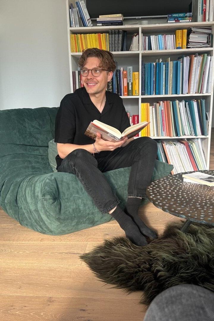Ein Mann sitzt lächelnd mit einem Buch auf einem grünen Sessel vor einem Bücherregal und einem runden Tisch.