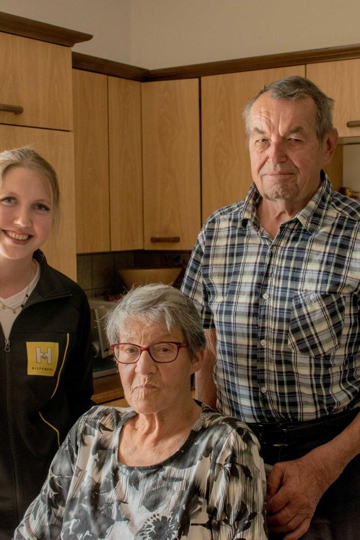 Eine junge Frau, ein älterer Mann und eine ältere Frau sitzen und stehen gemeinsam in einer Küche.