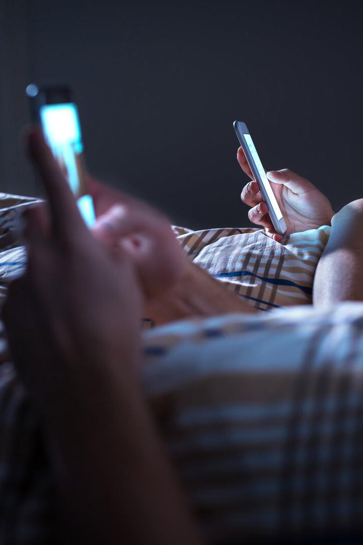 Zwei Personen liegen nebeneinander im Bett und benutzen jeweils ein Smartphone im Dunkeln.