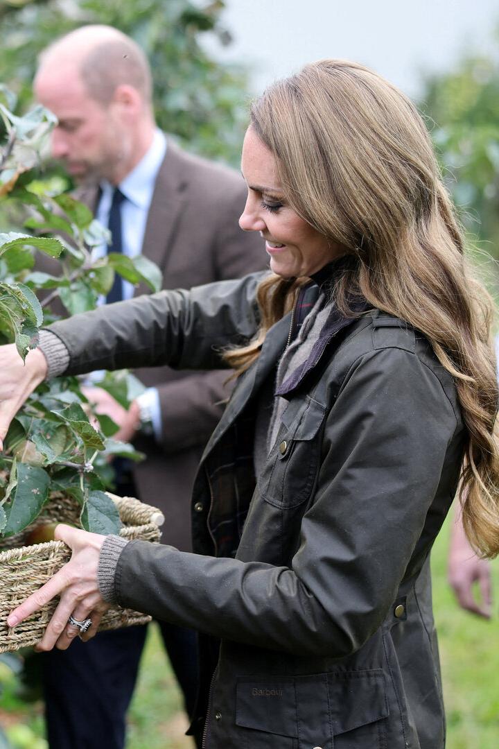 Kate pflückt Äpfel in einen Korb, während William im Hintergrund steht.