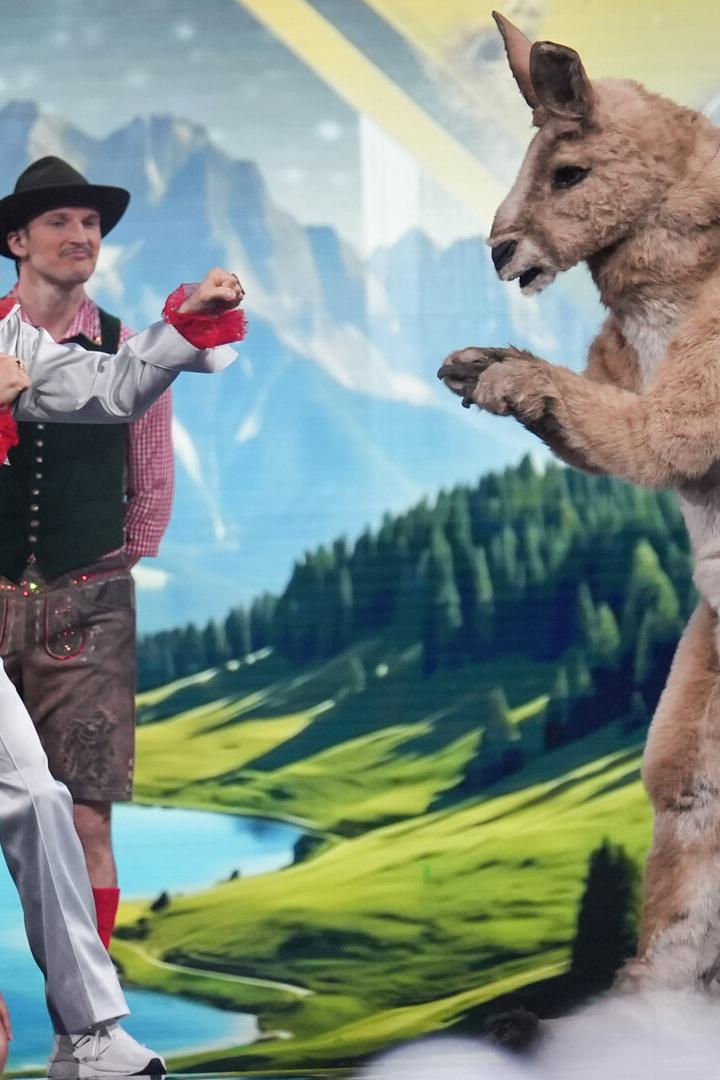 Mehrere Menschen in bunten Kostümen stehen vor einer Alpenlandschaft, einer davon kämpft spielerisch mit einem Känguru im Kostüm.