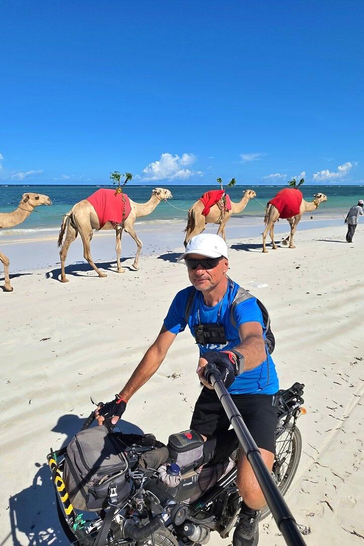 Ein Mann macht ein Selfie auf einem Fahrrad am Strand, während mehrere Kamele mit roten Decken am Meer entlanggeführt werden.